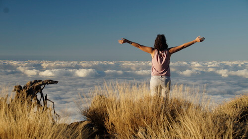 Das bedeutet Fesseln sprengen: Sandra steht mit erhobenen Händen über den Wolken auf einem Berg und schaut in die Weite
